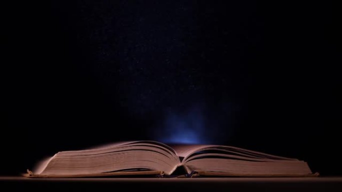 一本打开的书落在黑暗工作室的桌子上。探照灯发出的一束光照亮了它的书页和飞过它的尘埃颗粒。慢动作。特写