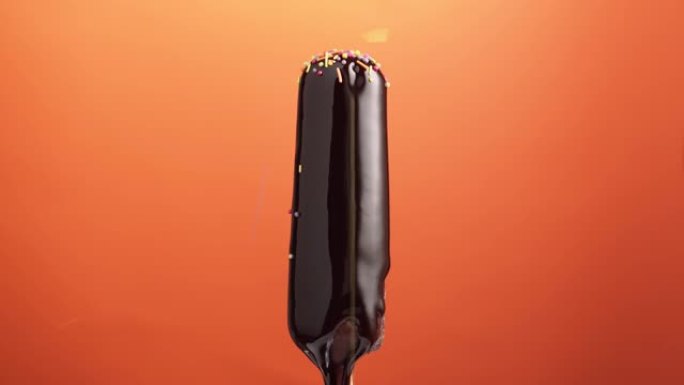 棍子上的冰淇淋，在橙色背景上涂有巧克力釉，旋转。五颜六色的洒落在巧克力浇头的冰棒上。