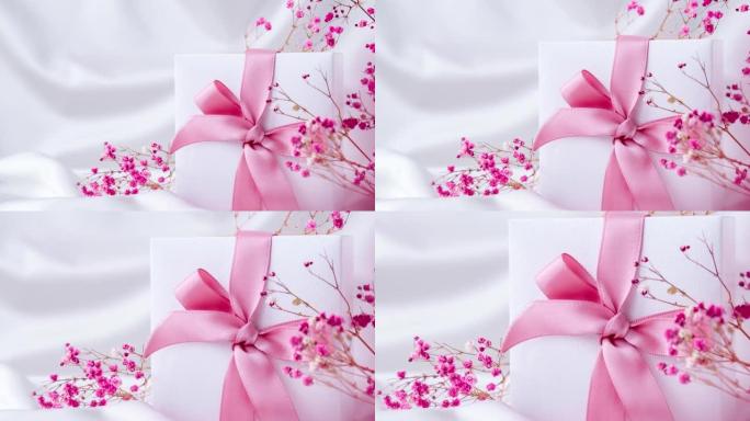 白色礼物盒，粉红丝带和白色丝绸织物背景上的粉红色小花。假日贺卡。4k视频。放大