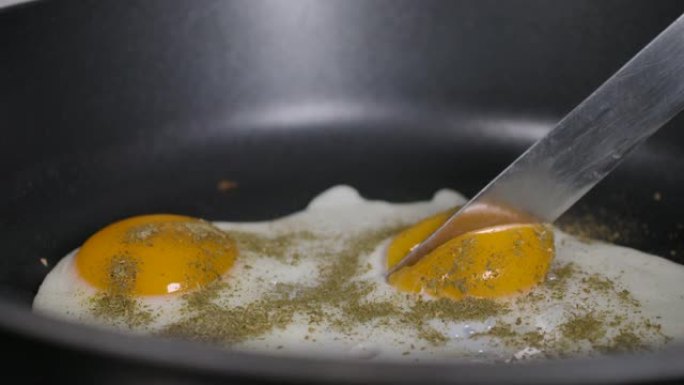 慢动作特写男人在煎锅上煮煎鸡蛋作为早餐，在家厨房。煎锅里的煎鸡蛋撒上磨碎的香料。厨师用刀慢动作切蛋黄