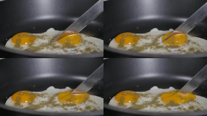 慢动作特写男人在煎锅上煮煎鸡蛋作为早餐，在家厨房。煎锅里的煎鸡蛋撒上磨碎的香料。厨师用刀慢动作切蛋黄