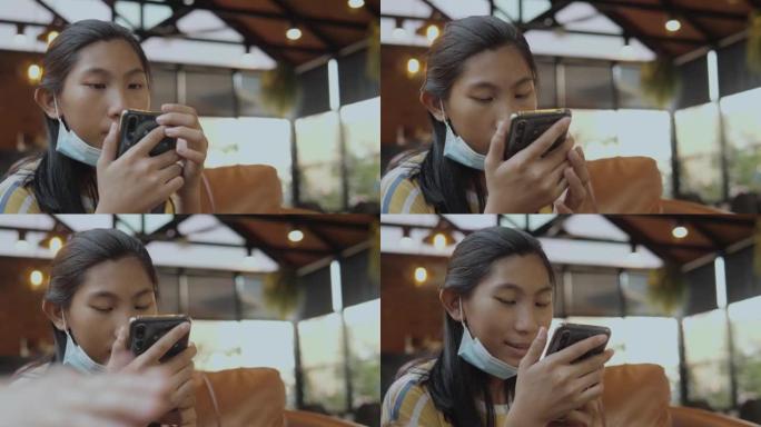 快乐的亚洲女孩戴着口罩，通过智能手机观看病毒视频，坐在沙发上与家人，生活方式的概念。