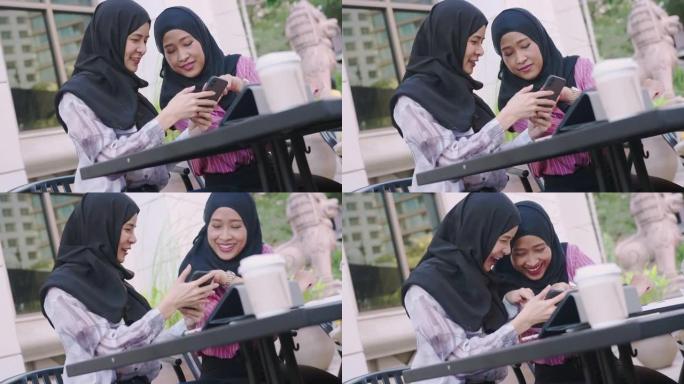 两名年轻的亚洲穆斯林女性戴着头巾坐在咖啡馆里，使用智能手机，购买新小工具，朋友见面约会，现代技术，在