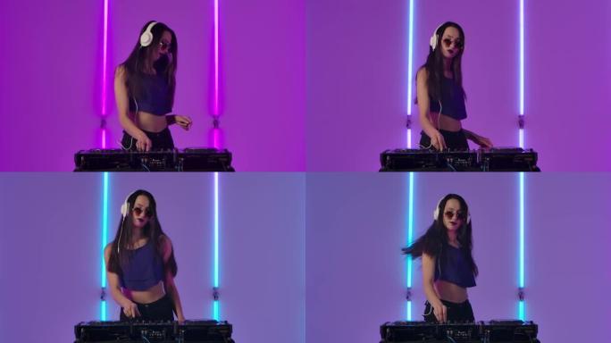 戴着太阳镜和大白色耳机的年轻时尚女性的肖像在DJ转盘上跳舞和混合音乐。时装模特在工作室明亮的霓虹灯背