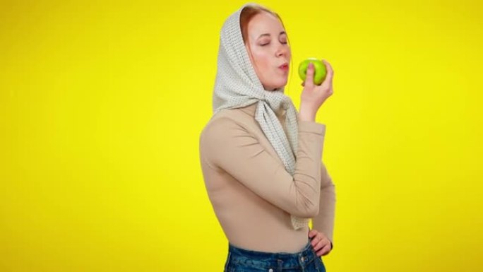 苗条的年轻女子站在黄色背景下咬着青苹果。自信的高加索女士穿着方巾享受美味水果的中景肖像看着相机微笑