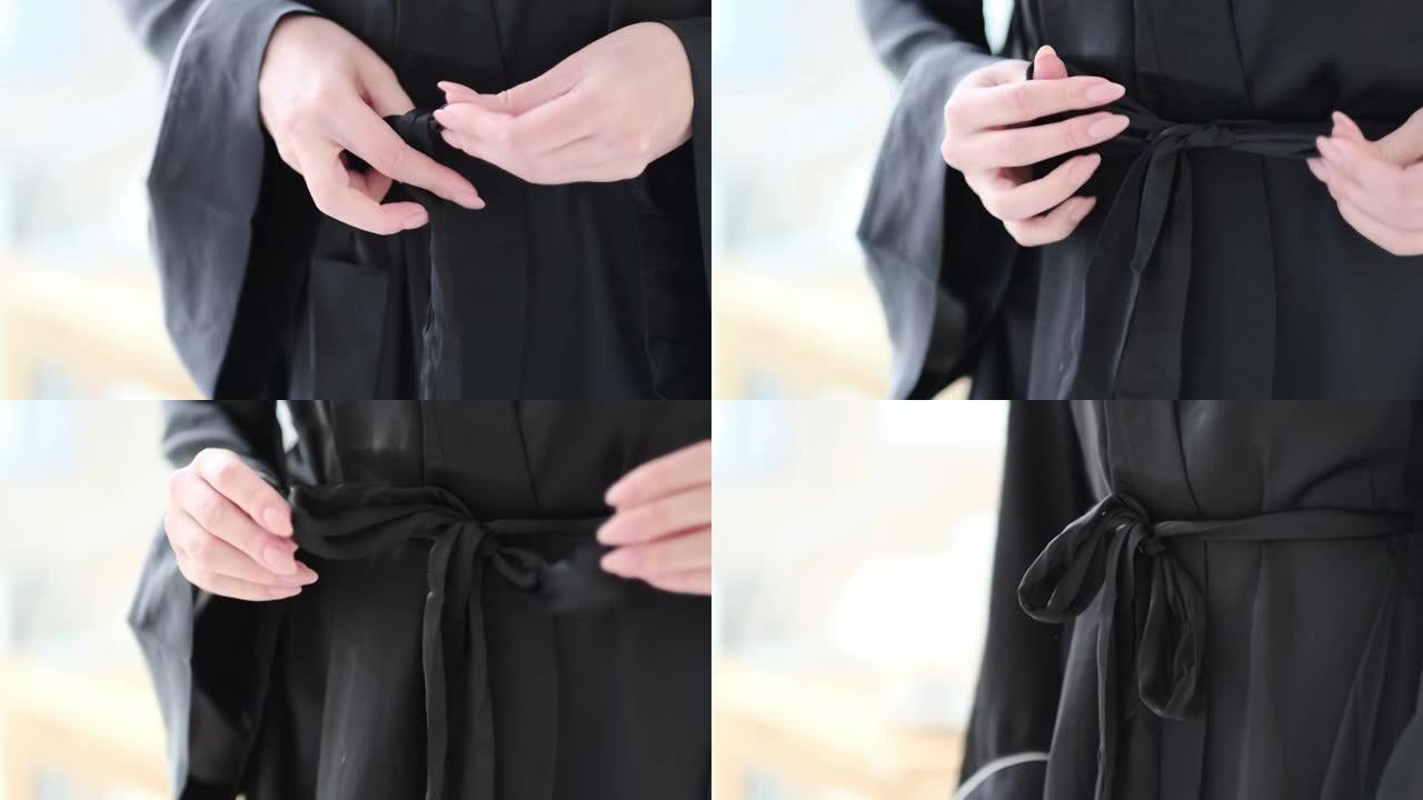 穿着黑色丝绸长袍的女人把腰带绑在长袍上。特写视图。慢动作视频。库存镜头