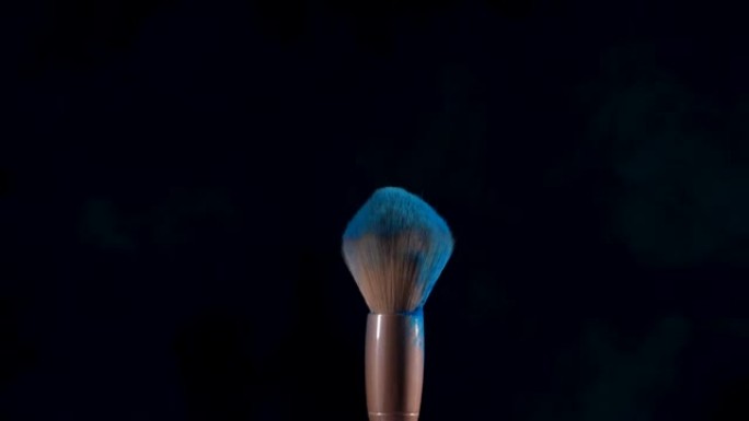化妆刷相互接触，蓝色压碎的油漆颗粒在黑色背景下飞入太空。美容行业，装饰化妆品概念。特写。慢动作