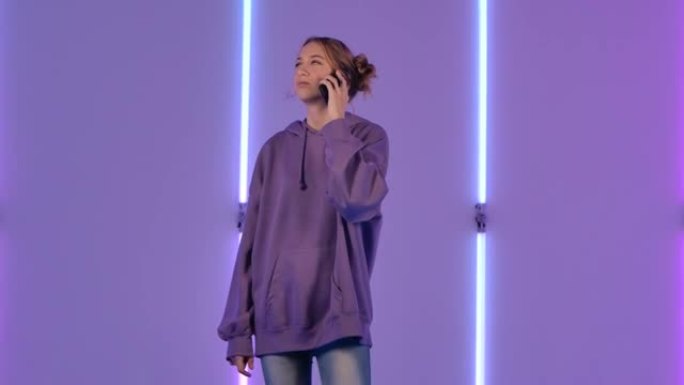 年轻迷人的女人为手机聊天并欢欣鼓舞的肖像。穿着紫色连帽衫的时装模特在黑暗工作室明亮的霓虹灯背景下摆姿