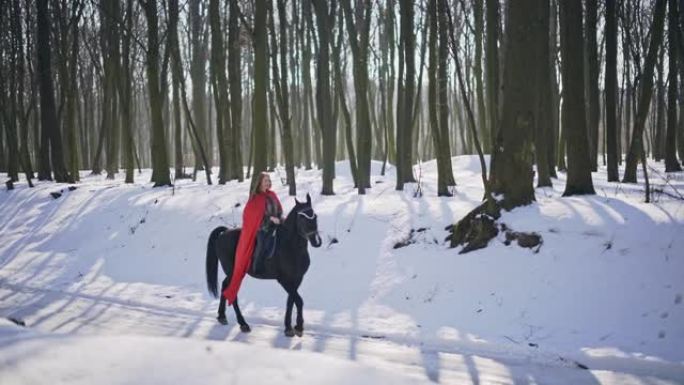 穿着红色斗篷的女人骑着马沿着白雪覆盖的道路穿过森林，冬天在白色的寒冷景观中