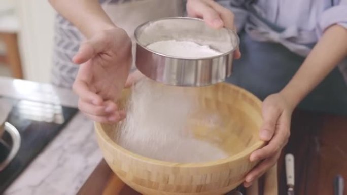 亚洲夫妇的亲密手互相帮助，在家里的厨房里，在浪漫的氛围中制作面包店。年轻人一起工作，将配料混合在木碗