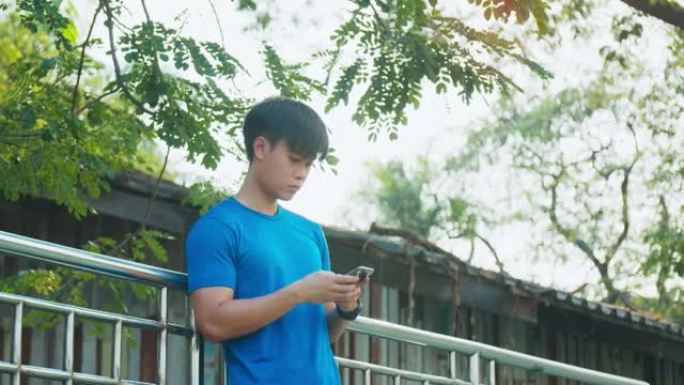 运动男子在跑步后站在公园的围栏上，使用智能手机检查社交媒体并在智能手机上发短信给朋友。