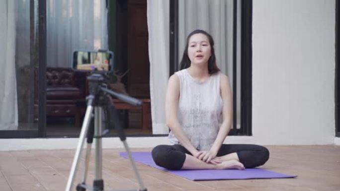 亚洲女性在线教练瑜伽演示通过移动智能手机三脚架直播向观众展示姿势。女性健身教程。培训师与在家在线教授