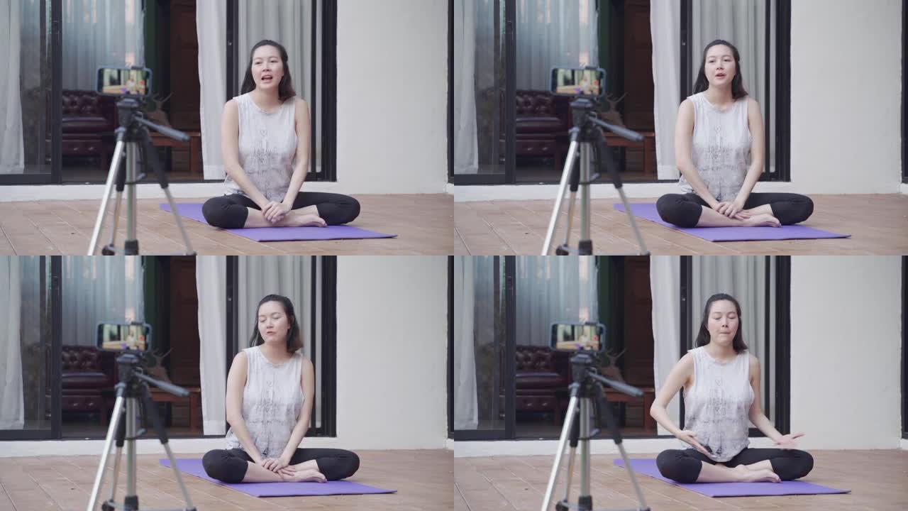 亚洲女性在线教练瑜伽演示通过移动智能手机三脚架直播向观众展示姿势。女性健身教程。培训师与在家在线教授