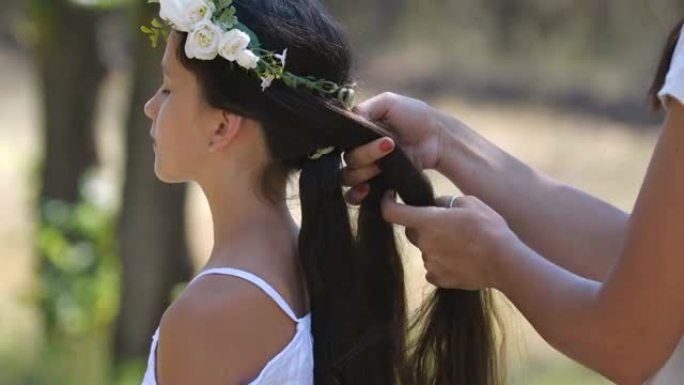 母亲照顾戴着白花花环的少女的长发。一名妇女在大自然背景下在户外编织女儿的辫子。轮廓肖像慢动作4k 5