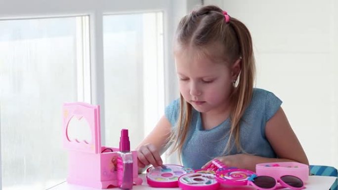 一个美丽的小女孩用蓝色的孩子画她的眼睛是阴影，照镜子。孩子在玩具美容院在家玩耍。