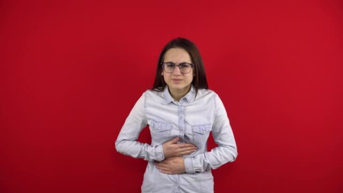 一个戴眼镜的年轻女子胃痛，她将其握在手中。在红色背景上拍摄。