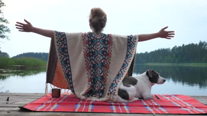 女人穿着舒适的雨披，双手伸开，欣赏大自然，欣赏日落时与白色和棕色的狗在湖面上的时光。自我护理概念