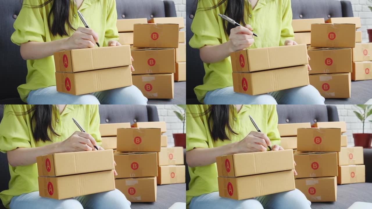亚洲年轻女性的特写手在家里的包裹箱上写下地址，用于向客户，运输和物流，在线商家和卖家，企业主或中小企