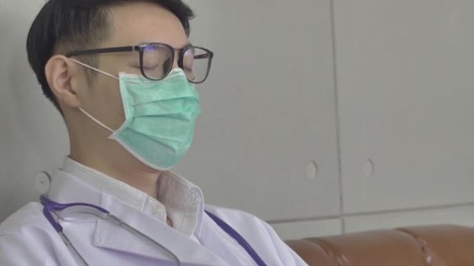 手持照片中，一位年轻医生戴着医用口罩和眼镜的工作精疲力尽，坐在沙发上。