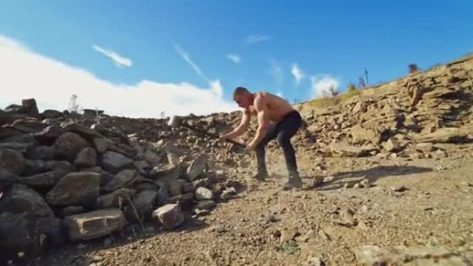 户外有巨大锤子的赤膊运动员。强壮的运动员在岩石山背景上用坚硬的钢锤打碎石头。