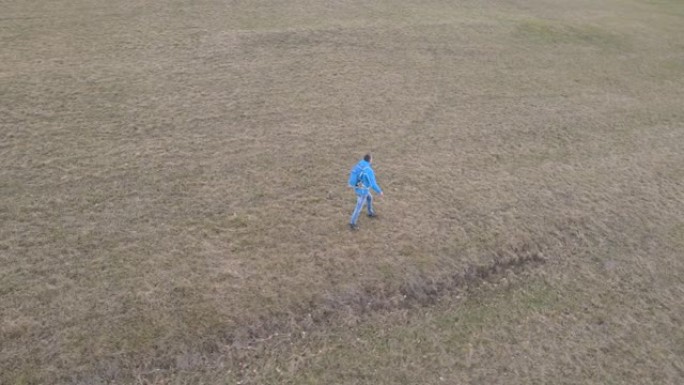 穿着蓝色夹克和蓝色背包的男性徒步旅行者穿过草地，被拍摄下来，随后是无人机
