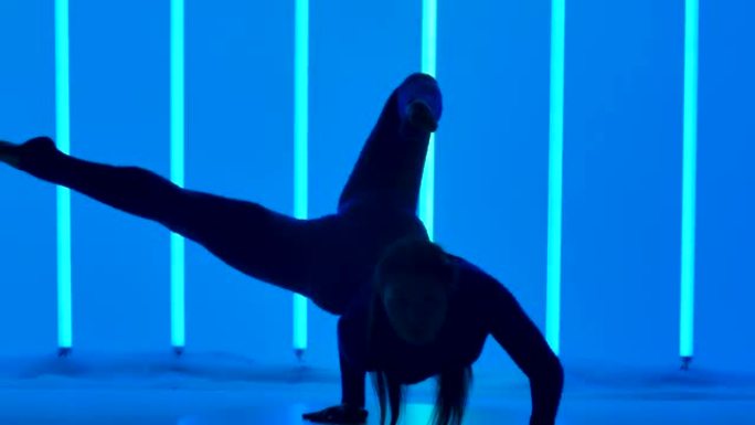 一个有魅力的女人表演倒立，移动她的腿，把它们分成一个裂口。工作室里的体操运动员在五彩霓虹灯的背景下。