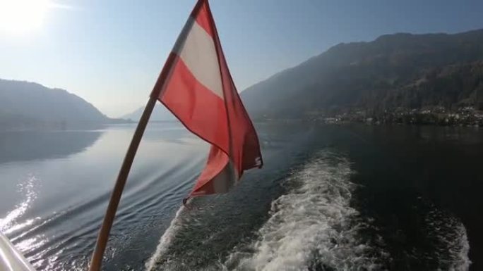 奥地利国旗安装在船轨的民意测验上。旗帜在风中轻轻地挥舞着。湖面四面环绕着高山，宁静而平静。乘船游览
