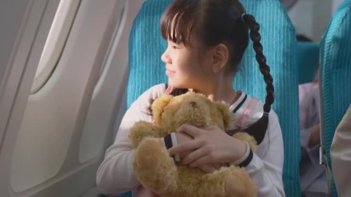 亚洲年轻小女孩拥抱熊娃娃，坐在飞机上的肖像。这个孩子和家人一起旅行感到兴奋和快乐。乘客在飞机上的运输