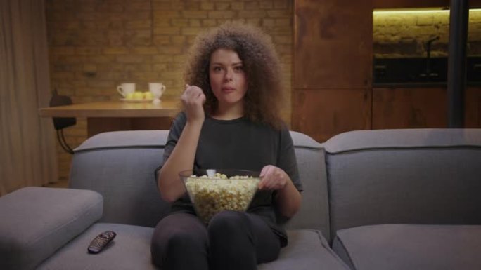20多岁的漂亮卷发女人看有趣的电视节目，坐在沙发上吃爆米花。微笑的女人喜欢独自在家看电影。