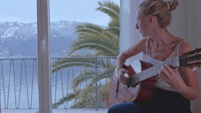 年轻女性在家弹吉他。窗外的湖泊和山脉景色