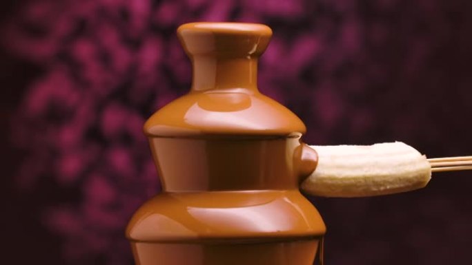 融化的牛奶巧克力在巧克力喷泉中流动。将成熟的香蕉浸入并包裹在从叶栅流下来的热液体巧克力中。火锅。派对