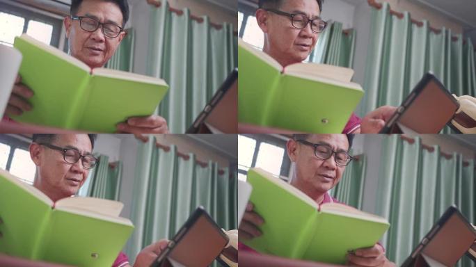 亚洲老人戴眼镜看书在家客厅书桌上敲击平板电脑，看书时专注，老年学习，阅读故事放松，中年业余爱好
