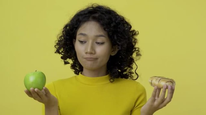 年轻漂亮的女人拿着甜甜圈和苹果在黄色背景。