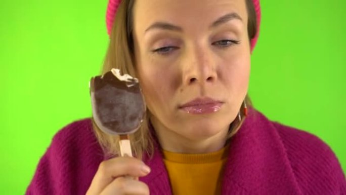 女人的肖像在巧克力釉的棍子上咬冰淇淋，然后冷冻，她变冷了。女人穿着针织帽，在工作室里穿着绿色背景的红