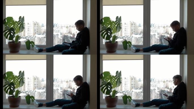穿着黑色连帽夹克的悲伤少年坐在窗台上，向窗外望去，并使用手机进行社交媒体，观看视频。在家中大流行期间