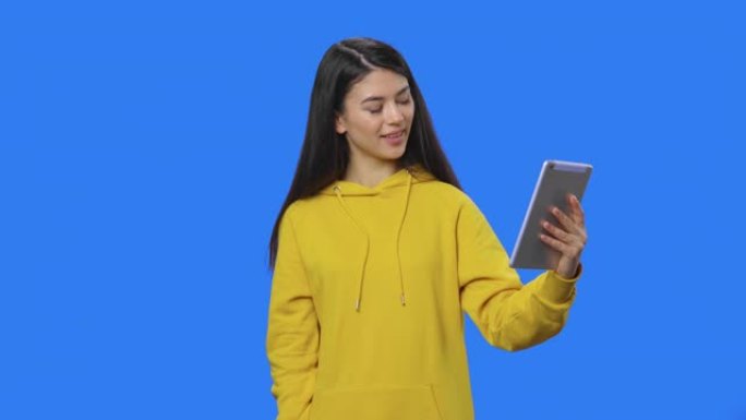 漂亮黑发女人使用平板电脑在视频通话中交谈的肖像。穿着黄色运动衫的长发年轻女子在蓝屏背景的工作室摆姿势