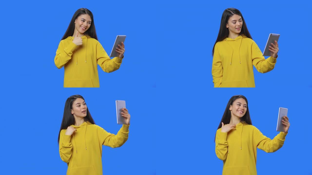 漂亮黑发女人使用平板电脑在视频通话中交谈的肖像。穿着黄色运动衫的长发年轻女子在蓝屏背景的工作室摆姿势