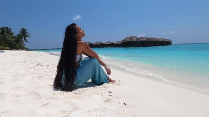 4k视频: 在马尔代夫的暑假期间，坐在沙滩上的亚洲妇女在沙滩上玩乐并享受阳光