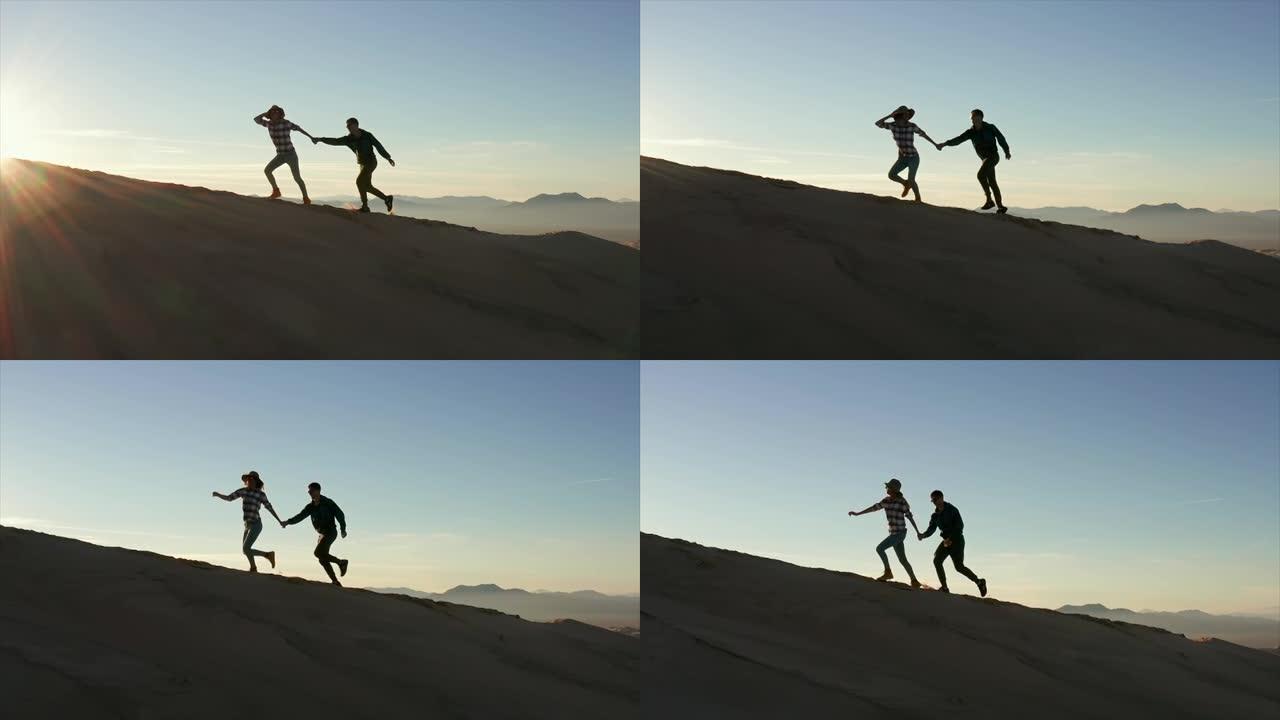 慢动作无人机射击，一对夫妇在日落时分在发送沙丘的边缘奔跑。旅行和生活方式概念