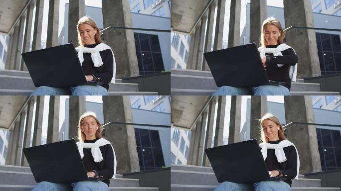 漂亮的平静微笑的肖像20岁的浅色女孩穿着休闲服，坐在建筑物的台阶上，在笔记本电脑上工作，并在手持时钟