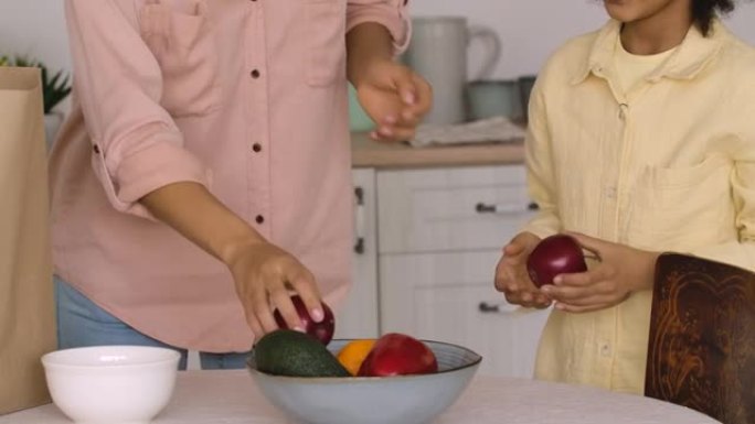 非裔美国妇女和女孩从纸袋中取出物品，将蔬菜和水果放在盘子上。母女俩在明亮的厨房摆姿势。特写。慢动作准