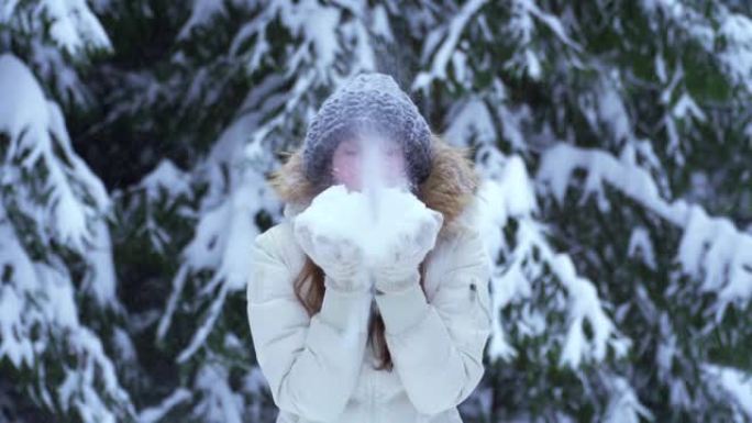 女人从手掌吹雪。把雪吹进相机里。冬天，在树木间的白雪皑皑的森林里，雪球在一个美丽的高加索女人手中。飘