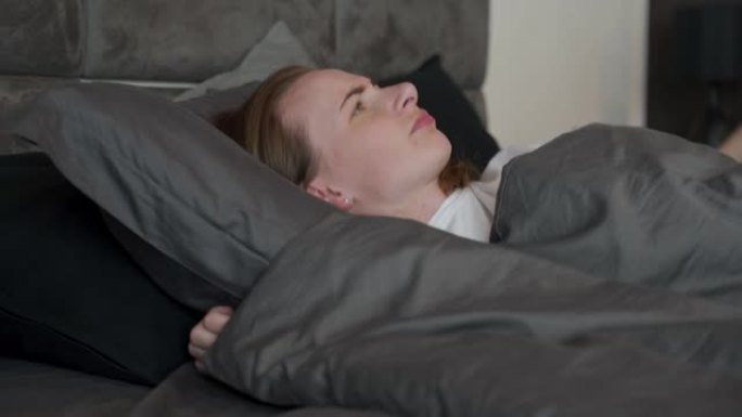 疲惫的女人睡不着。一个失眠的女人用枕头遮住耳朵。由于邻居吵闹，我半夜在床上醒来