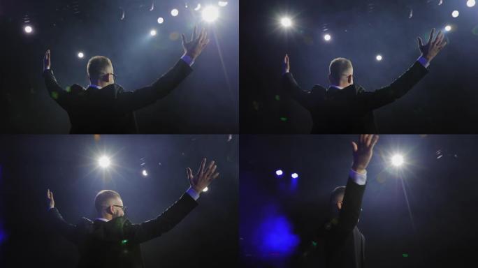 一个穿着西装的男人站在舞台上。聚光灯照在他身上。他举起双手大笑。演讲者的演讲。4K。