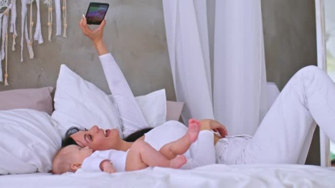 美丽的妈妈和婴儿躺在床上，用手机拍摄视频，家庭价值观。年轻的母亲在智能手机上录制视频，进行视频通话。