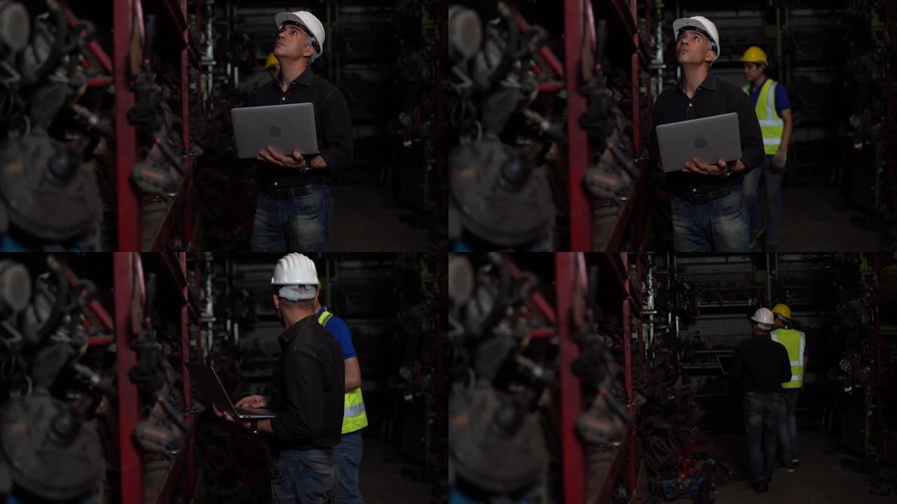 高级工程师使用笔记本电脑检查清单旧二手车发动机与年轻的亚洲工人在汽车配件商店仓库。老经理在回收汽车厂