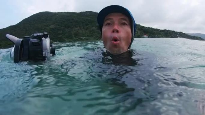 惊讶的冲浪摄影师。戴着蓝色帽子的男人带着动作相机在海里游泳，面容惊讶地环顾四周