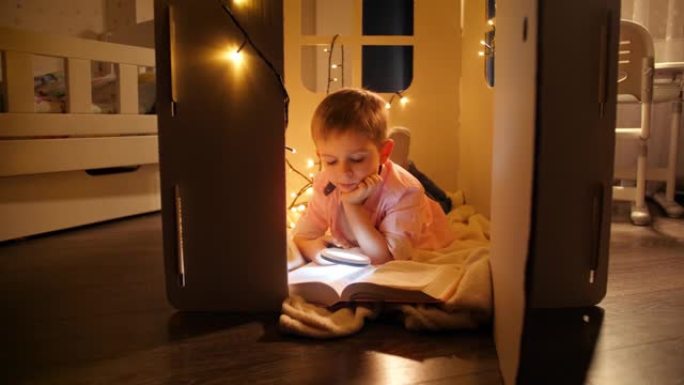晚上躺在玩具屋或帐篷里的男孩读书的多莉镜头。儿童教育与暗室阅读的概念