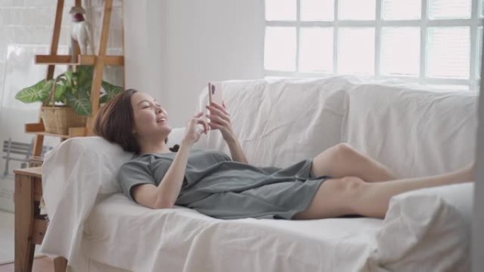 快乐的亚洲女人脸上放松使用手机上网和在家沙发上聊天。女人躺在客厅里，使用手机时尴尬地微笑。女人笑得开