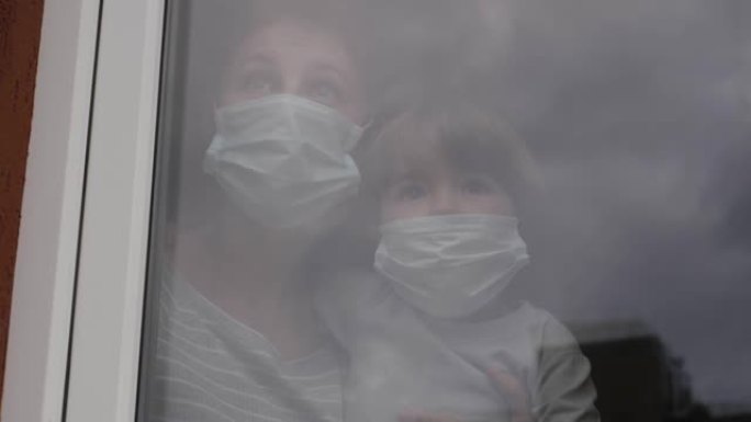 成熟的女人带着她的儿子，戴着防护面具，悲伤地透过窗户看着担心新型冠状病毒肺炎封锁。戴着保护面具的家庭